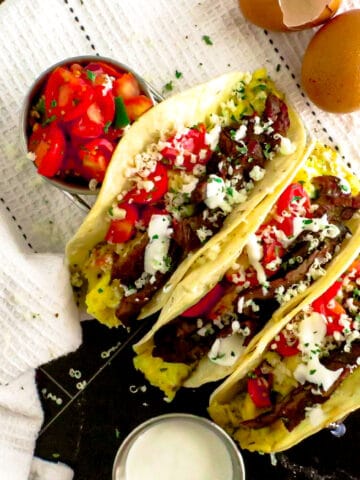 three tacos, filled with steak and scrambled eggs. Topped with tomatoes, crema and cheese