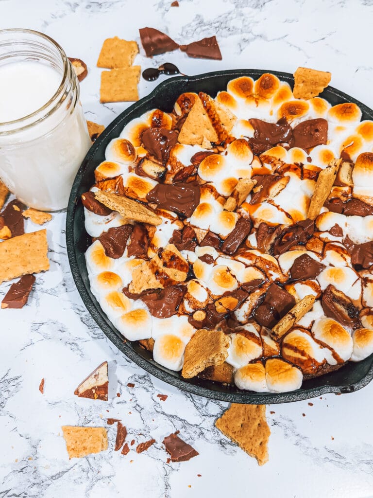 A glass of milk next to a cast iron skillet s'more. The skillet s'more is topped with toasted marshmallows, twix, hershey's, graham crackers and chocolate drizzle.
